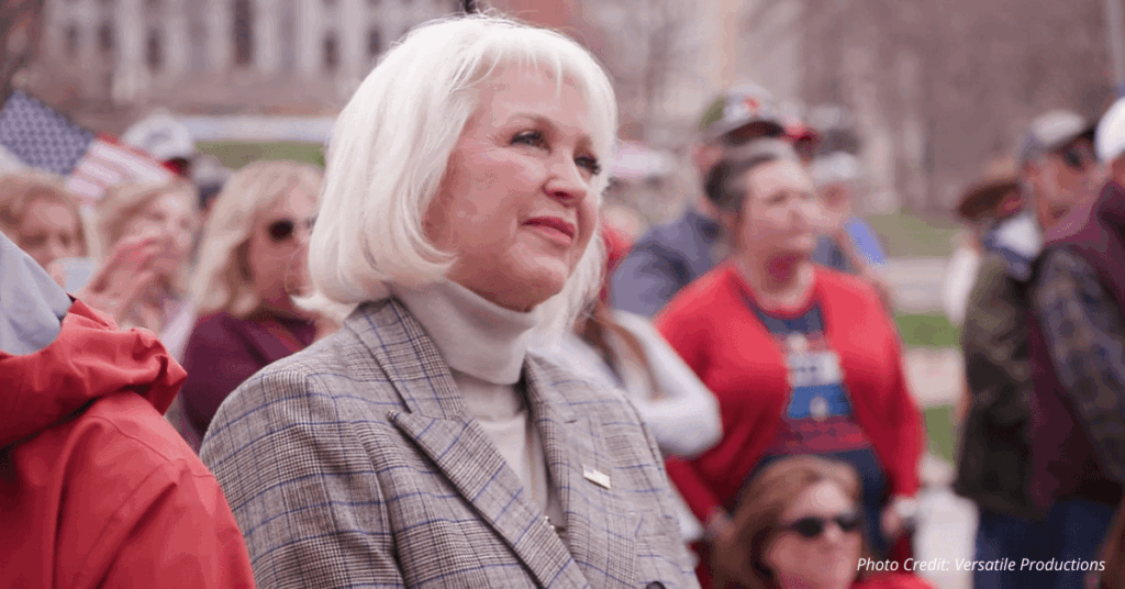 Tina Peters standing in a crowd at a public rally, looking ahead with a calm expression