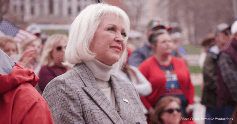 Tina Peters standing in a crowd at a public rally, looking ahead with a calm expression