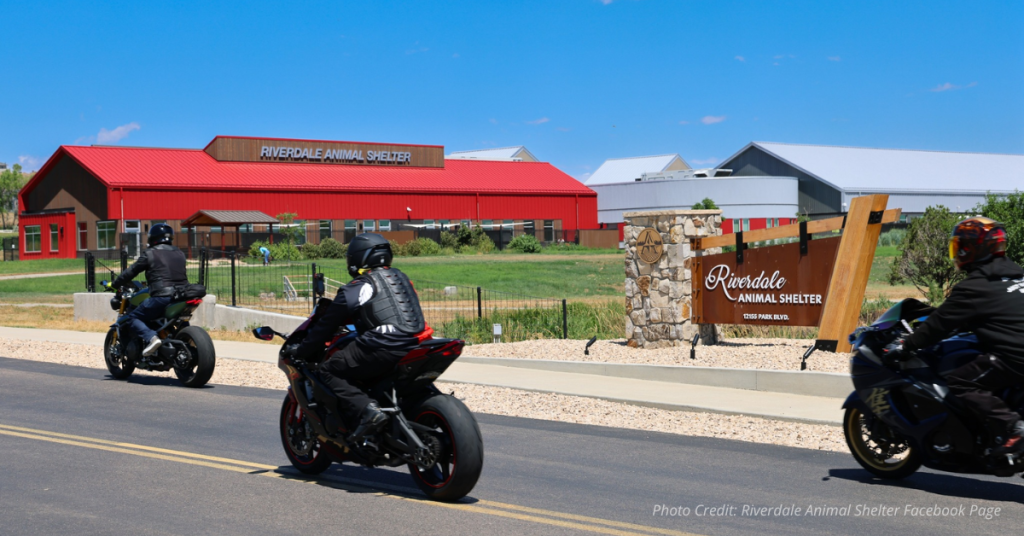 Riverdale Animal Shelter with sign and three motorcyclists passing on Park Blvd under a blue sky
