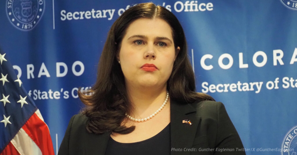 Jena Griswold standing at a Colorado Secretary of State press backdrop during a press conference