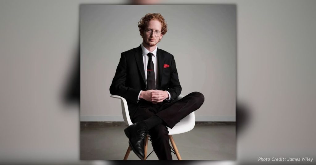 James Wiley, Colorado Secretary of State candidate, seated in a white chair wearing a black suit, black tie, glasses, and a red pocket square against a plain gray background