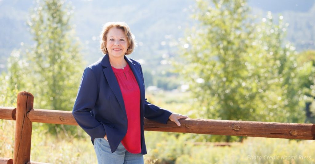 Naomi Riess, Republican candidate for Colorado House District 59, standing outdoors by a wooden fence with mountains and trees behind her