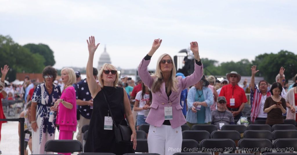 Two attendees raise their hands during Rededicate 250 on the National Mall, with the U.S. Capitol dome visible behind them, May 17, 2026