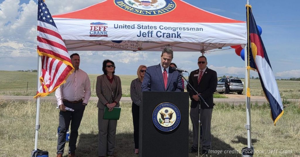 Rep. Jeff Crank speaks at an outdoor event beneath a congressional tent flanked by U.S. and Colorado flags.