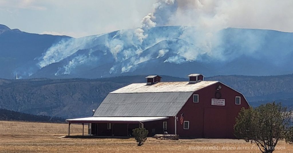 Aspen Acres Fire burns across Colorado mountains behind a barn as smoke rises from multiple areas.