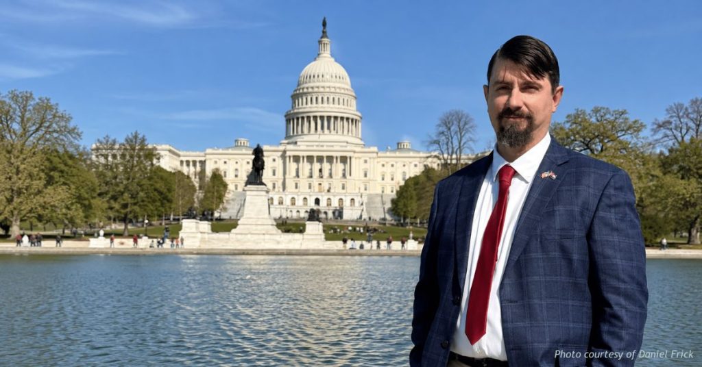 Daniel Frick stands in front of the U.S. Capitol dome and reflecting pool, wearing a blue suit and red tie.