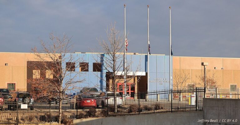 Exterior of the Aurora ICE Processing Center in Colorado, showing the blue entrance, parking lot, security fencing and flags at half-staff.