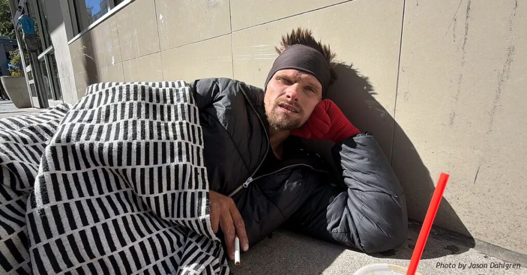 A man rests against a building wall on a Denver sidewalk, wrapped in a black-and-white patterned blanket and wearing a dark jacket.