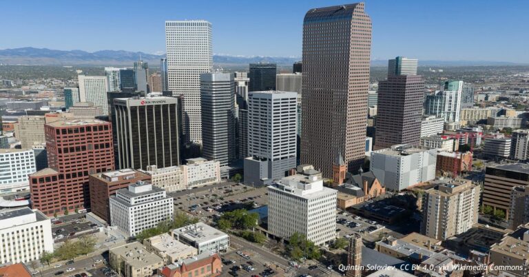 Aerial view of downtown Denver's office towers and surrounding buildings, with the Rocky Mountains in the distance.